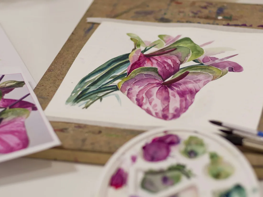 Lámina de acuarela botánica en proceso sobre la mesa de trabajo, con una flor de anturio pintada en rosa y verde, paleta con pigmentos y pinceles al lado